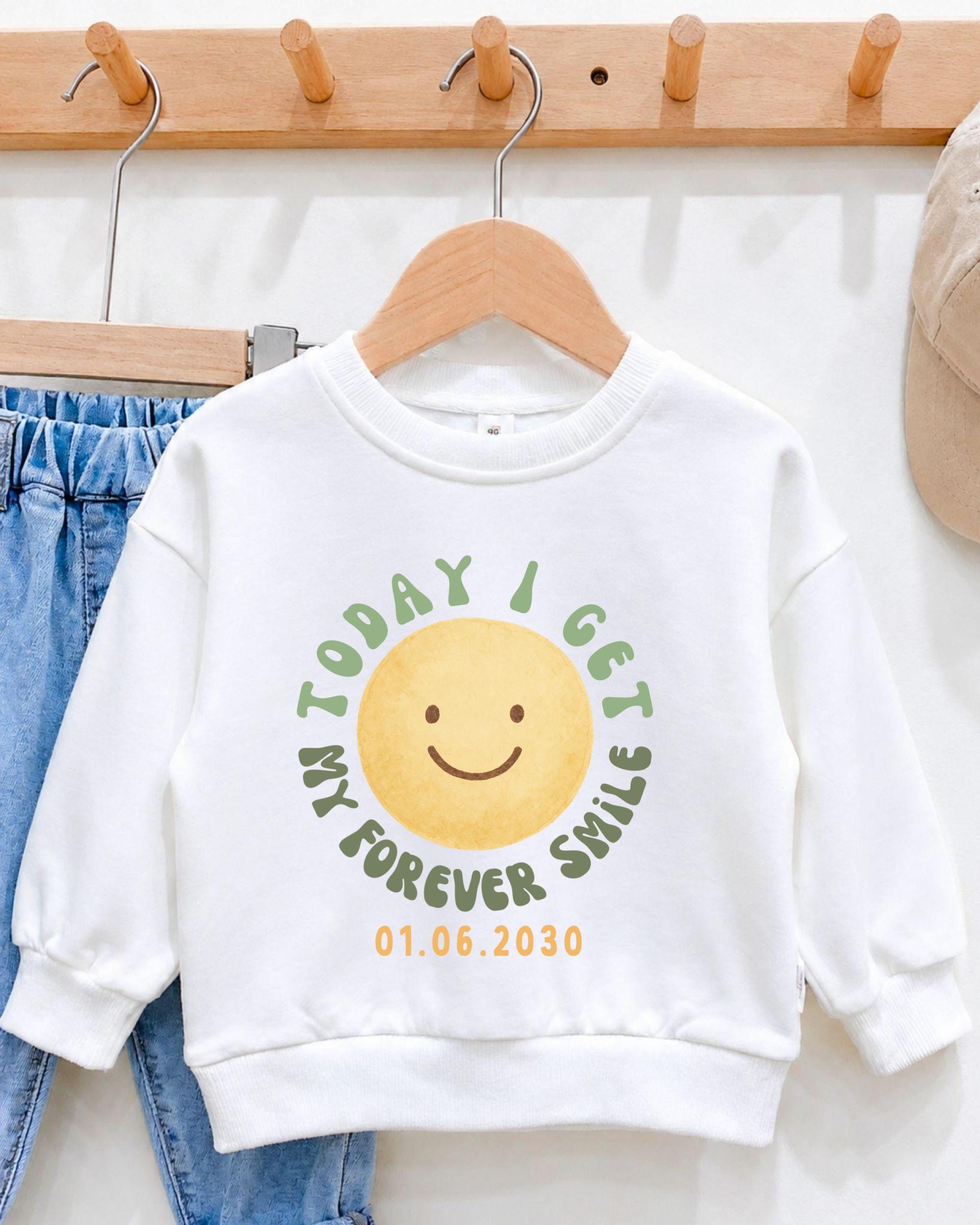 A white sweatshirt with a graphic design featuring a smiling yellow smiley face and the text "Today I get my forever smile" printed on it.