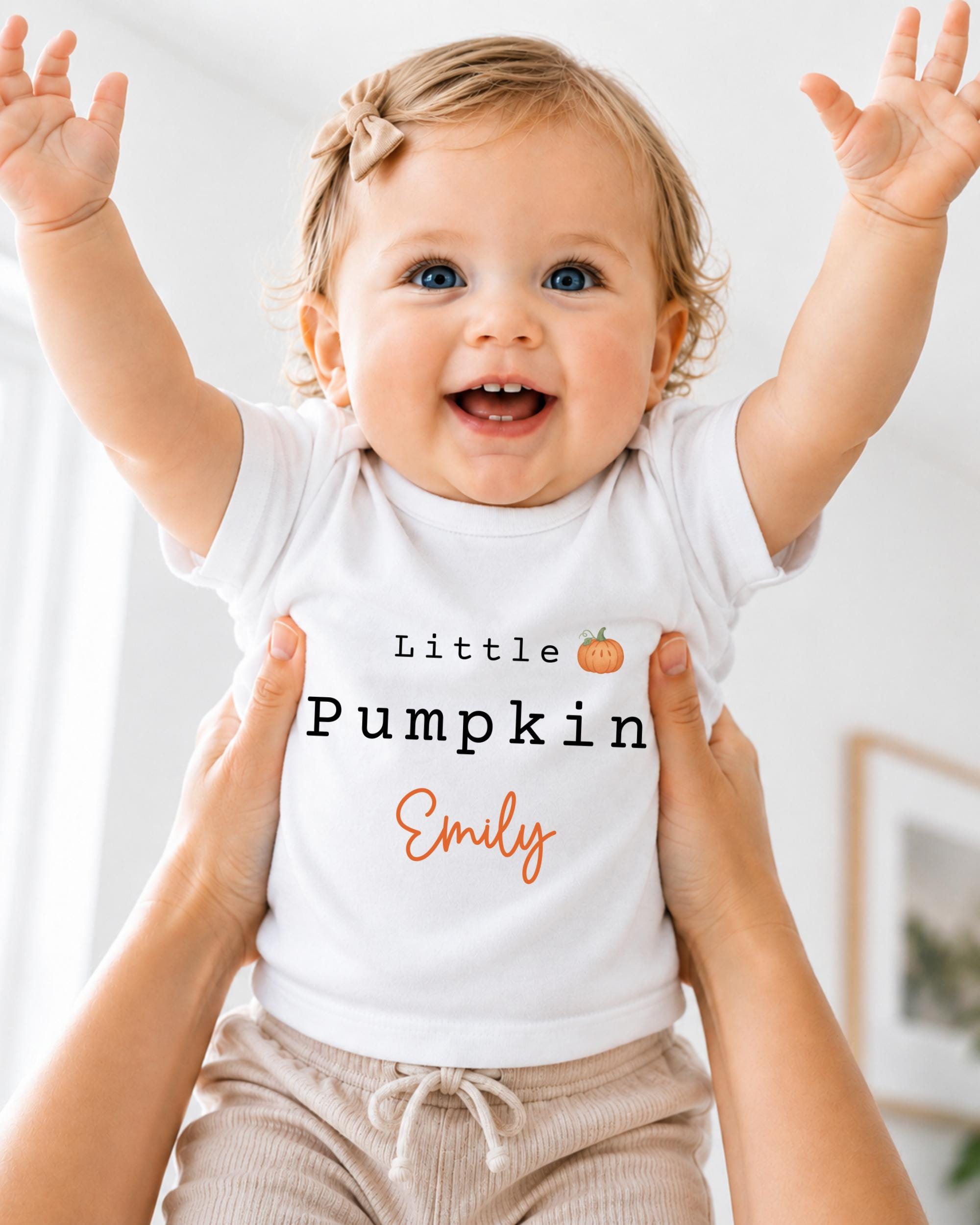 A smiling baby girl wearing a white t-shirt with the text "Little Pumpkin Emily" is being held up by someone's arms.