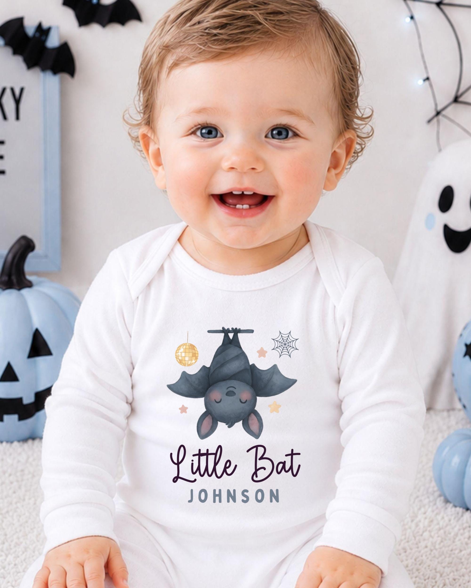 A smiling baby boy wearing a white onesie with a bat and spider design, sitting in front of Halloween decorations.
