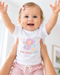 A smiling baby girl wearing a white t-shirt with a mermaid graphic and the text "Birthday Girl Jessica" is being held up by someone's arms.