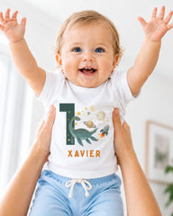 A smiling baby wearing a white t-shirt with a dinosaur and space-themed design, being held up by someone's arms.