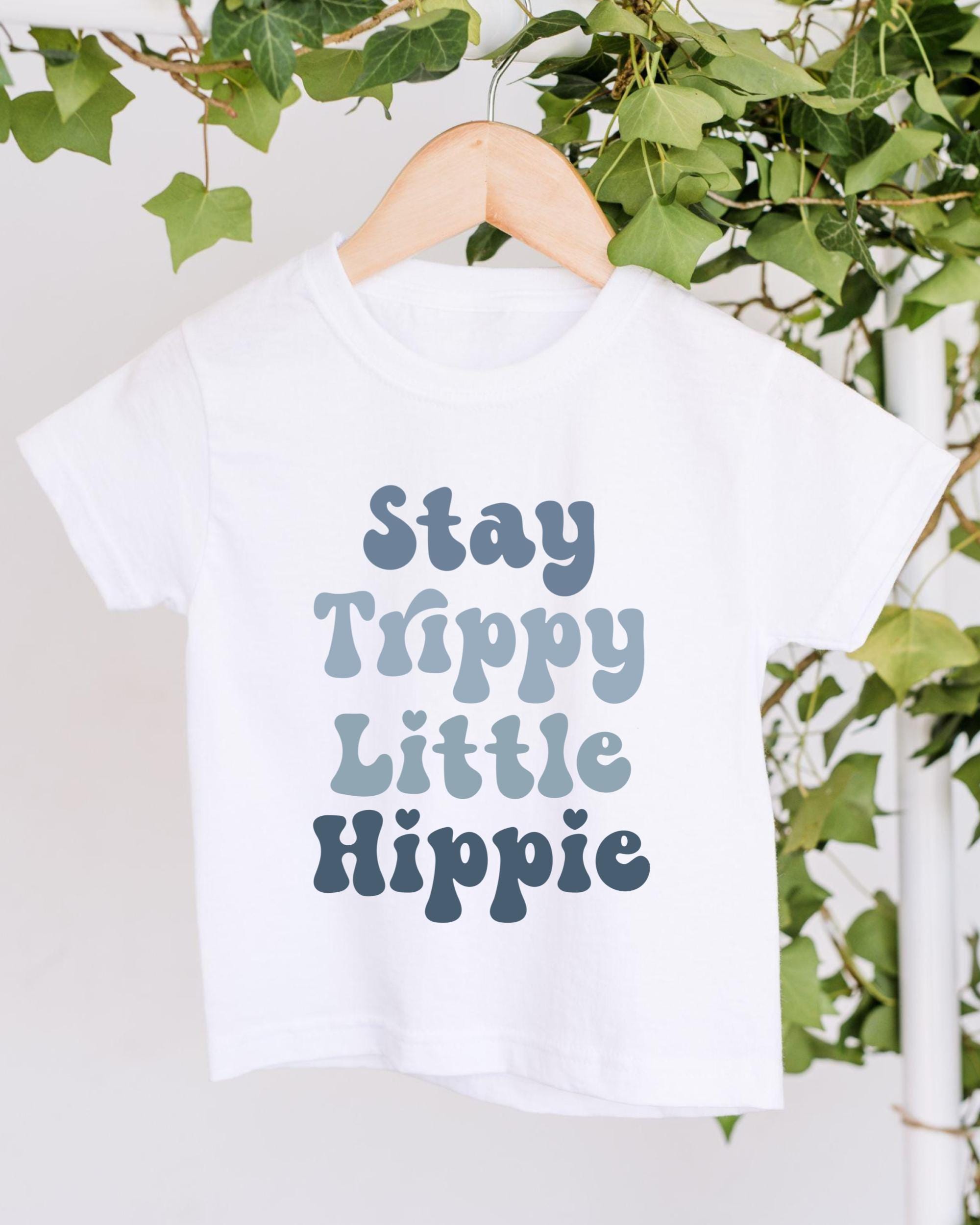A white t-shirt with the text "Stay Trippy Little Hippie" printed on it, hanging on a wooden hanger.