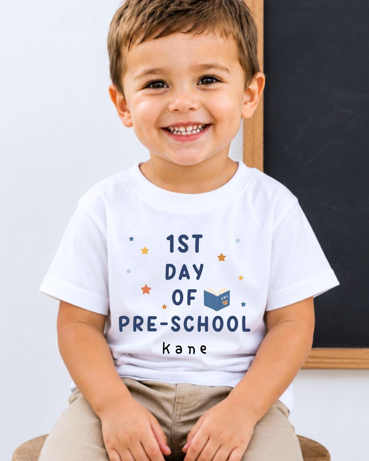 A young boy wearing a white t-shirt with the text "1st DAY OF PRE-SCHOOL kane" is smiling at the camera.