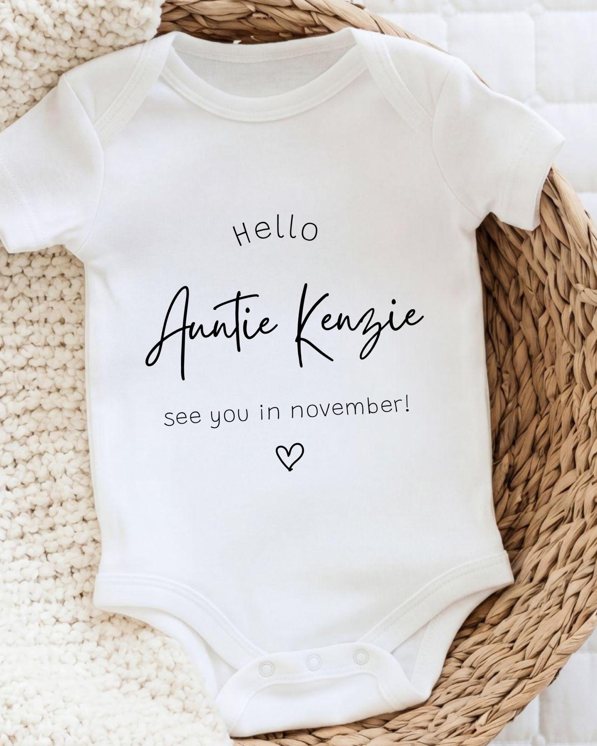 A white baby onesie with the text "Hello Auntie Kenzie see you in November!" printed on it, placed on a woven basket.