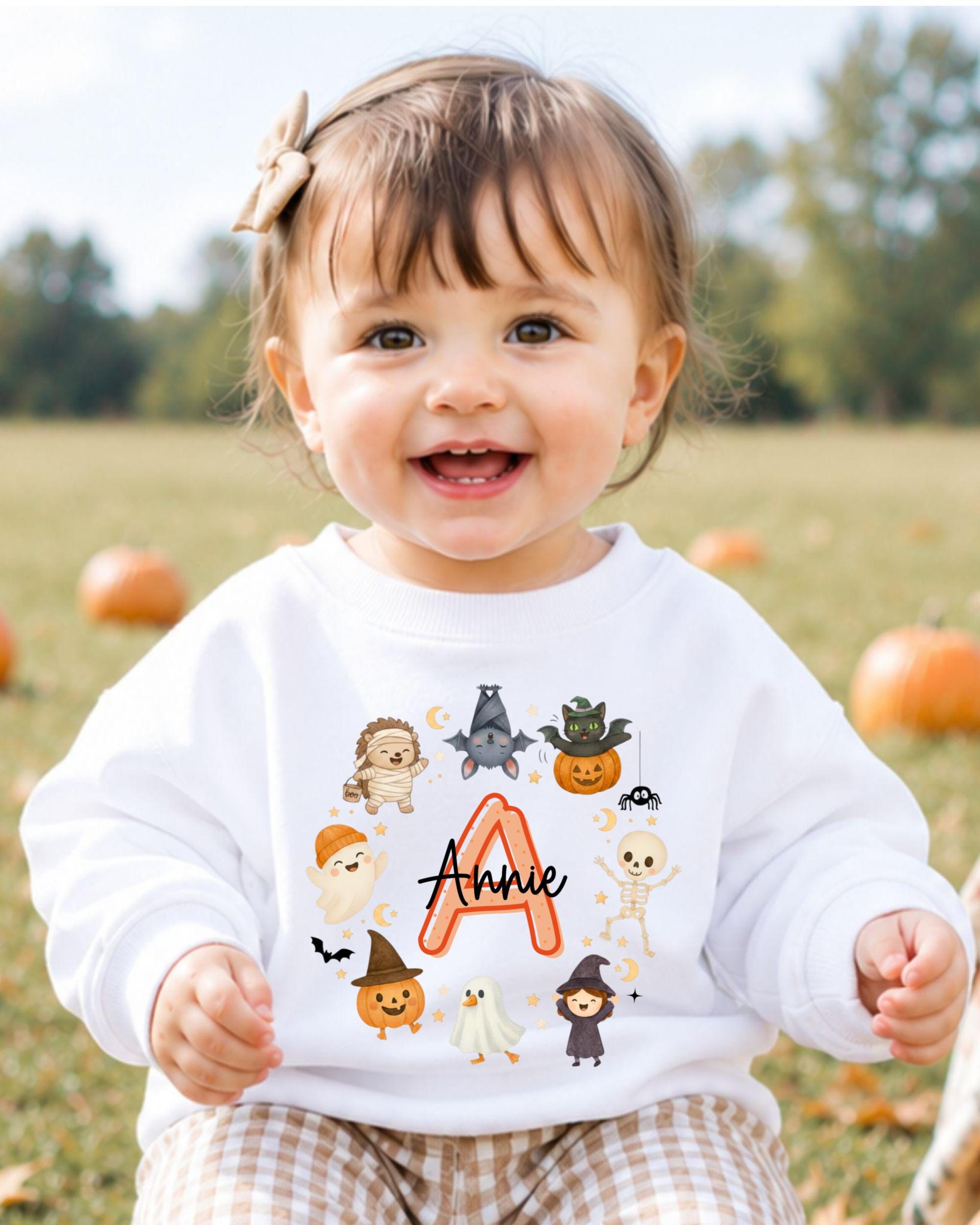 A smiling baby girl wearing a white sweater with a Halloween-themed design and the name "Annie" printed on it.