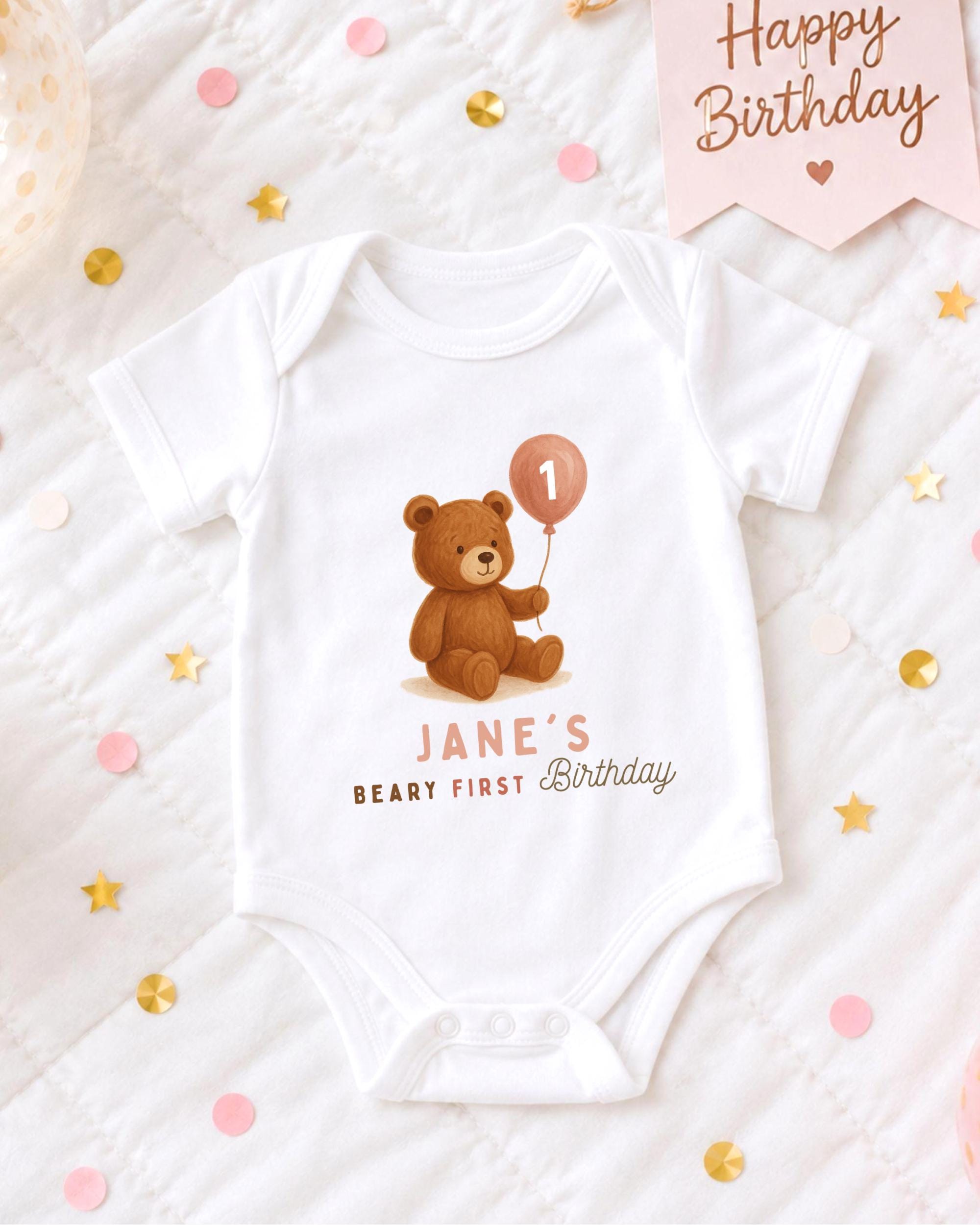 A white onesie with a teddy bear and a balloon is displayed on a bed with confetti.