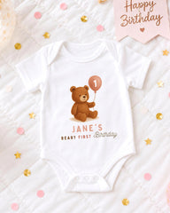 A white onesie with a teddy bear and a balloon is displayed on a bed with confetti.