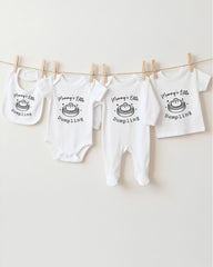A row of white baby clothes with the text "Mum's Little Dumping" printed on them, hanging on a clothesline.