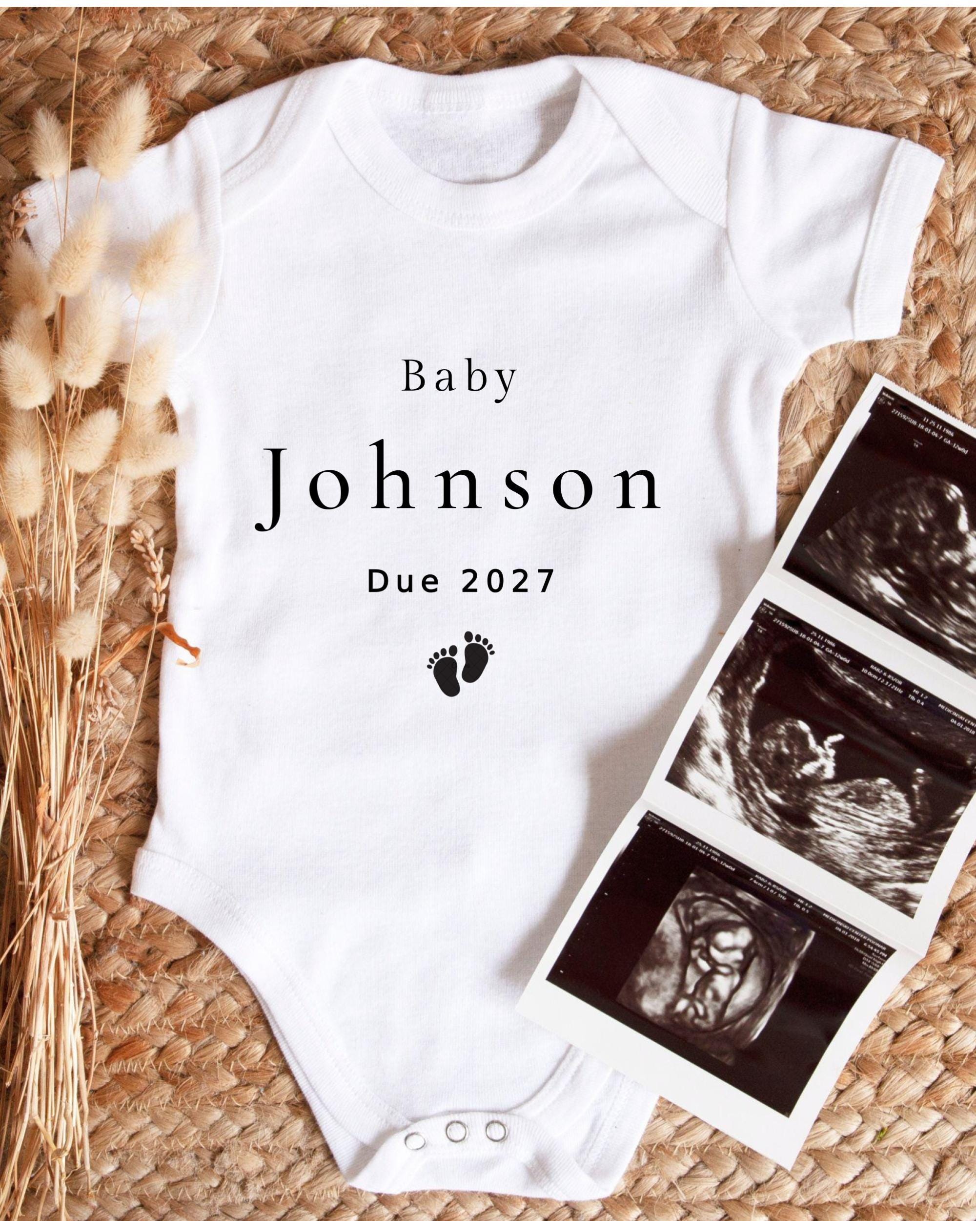 A white baby onesie with the text "Baby Johnson Due 2027" printed on it, along with a black and white image of an ultrasound scan.