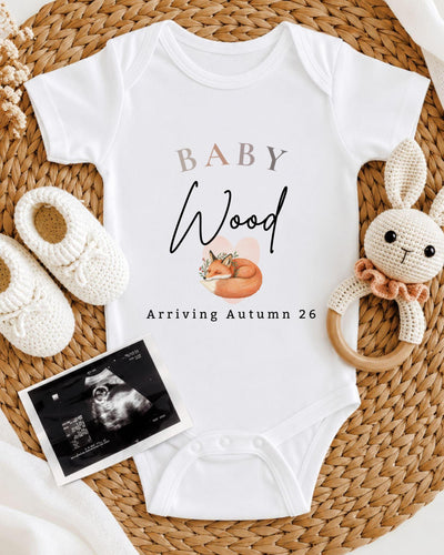 Personalisable white Baby Vest with beige 'BABY', black script 'Wood', a sleeping orange fox and 'Arriving Autumn 26' lettering.