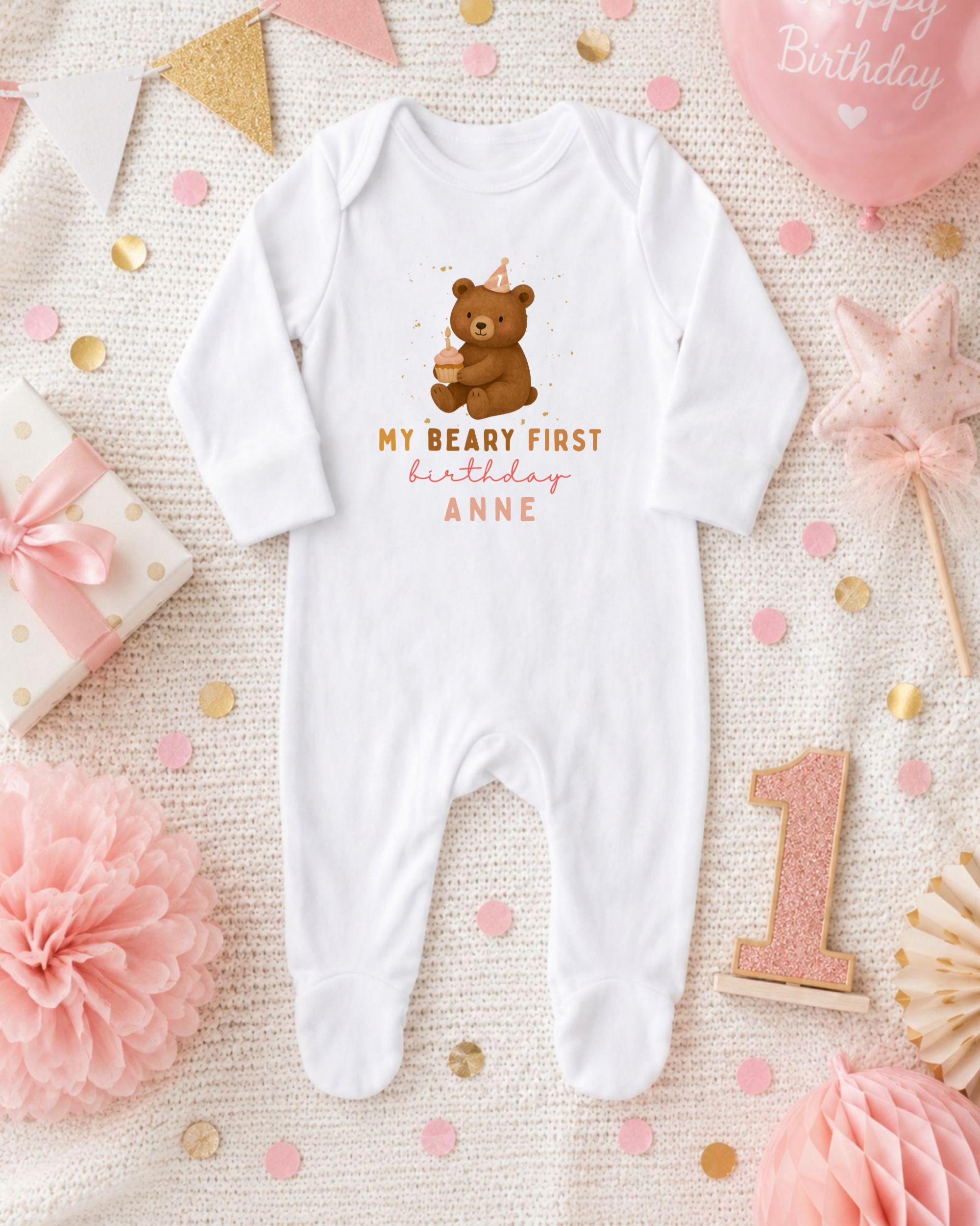 A white onesie with a teddy bear and the text "My Beary First birthday Anne" printed on it, surrounded by pink and gold confetti, a pink balloon, and other baby-related items.