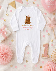 A white onesie with a teddy bear and the text "My Beary First birthday Anne" printed on it, surrounded by pink and gold confetti, a pink balloon, and other baby-related items.