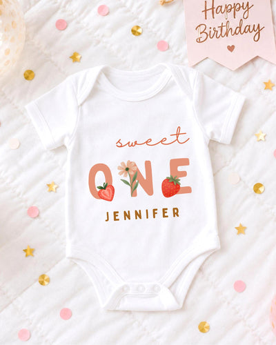 A white bodysuit with the text "Sweet One" printed on it, surrounded by confetti and a "Happy Birthday" sign.