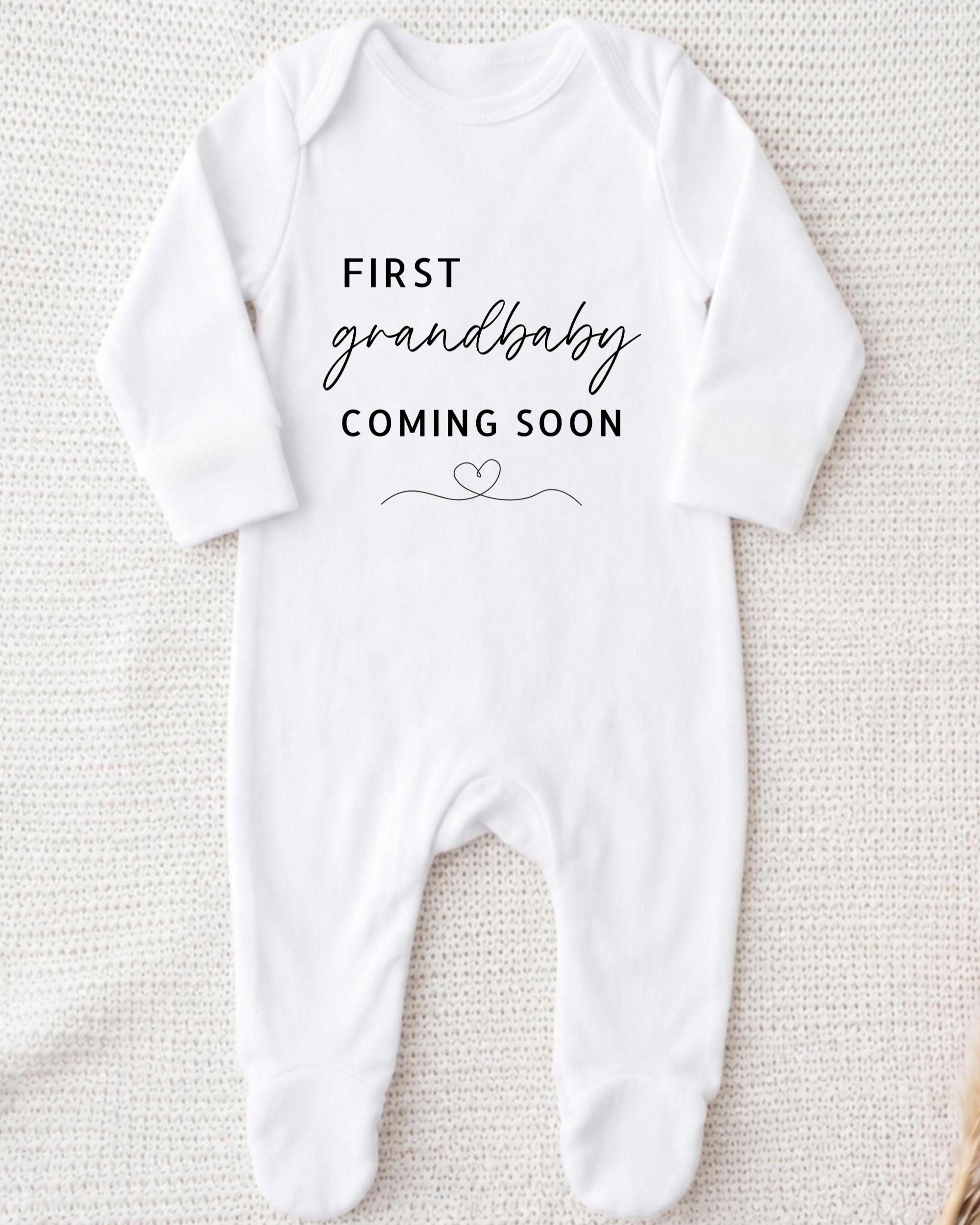 A white onesie with the text "FIRST GRANDBABY COMING SOON" printed on it.