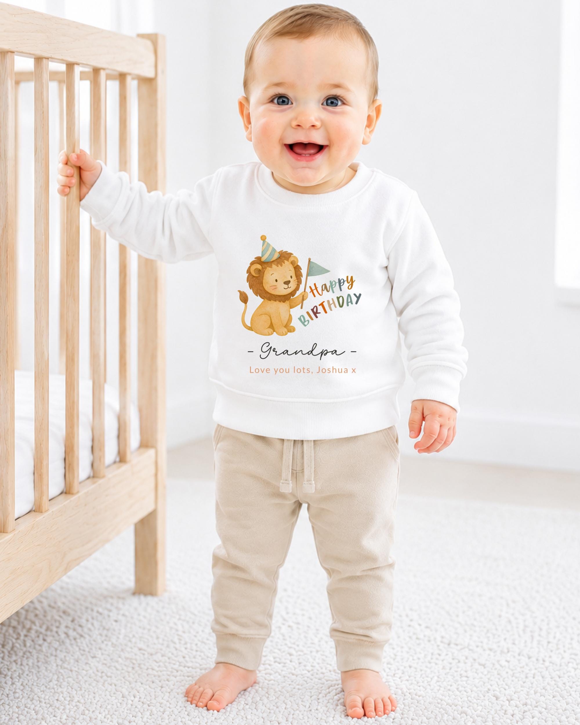 A smiling baby boy wearing a white sweatshirt with a cartoon lion and the text "Happy Birthday Grandpa" printed on it.