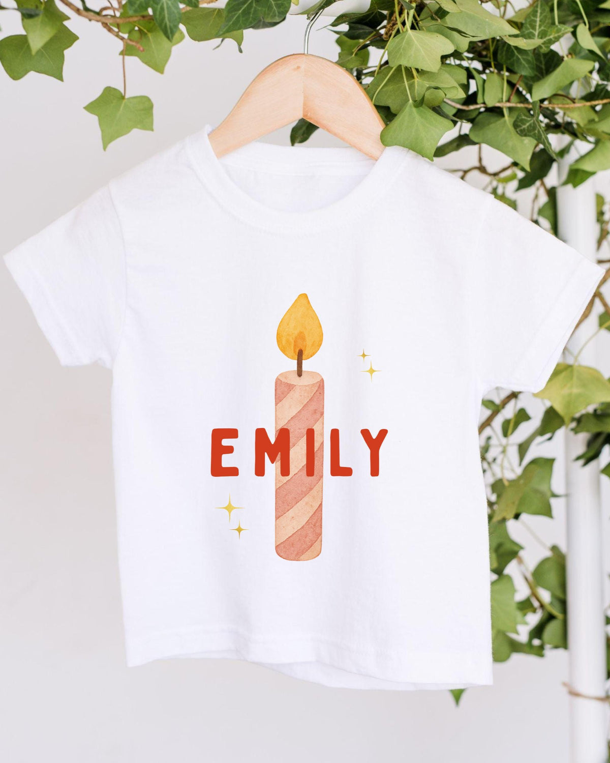 A white t-shirt with a graphic design of a lit candle and the name "EMILY" printed on it, hanging on a wooden hanger.