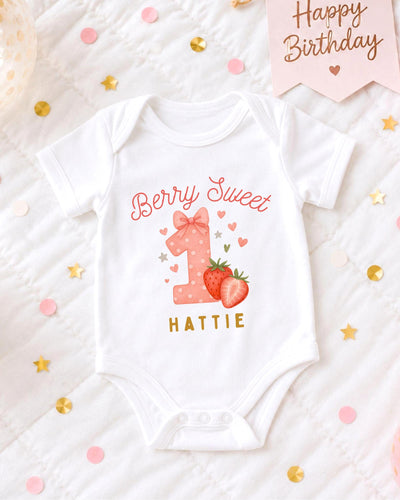 A white bodysuit with a pink bow and the text "Berry Sweet" printed on it, along with a strawberry and a heart, is displayed on a bed with confetti.