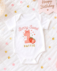 A white onesie with a pink bow and the text "Berry Sweet" printed on it, along with a strawberry and a heart, is displayed on a bed with confetti.