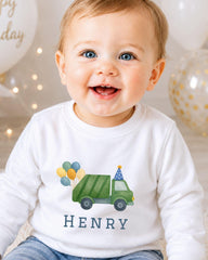 A smiling baby boy wearing a white shirt with a green truck and balloons printed on it, with the name "HENRY" written below the truck.