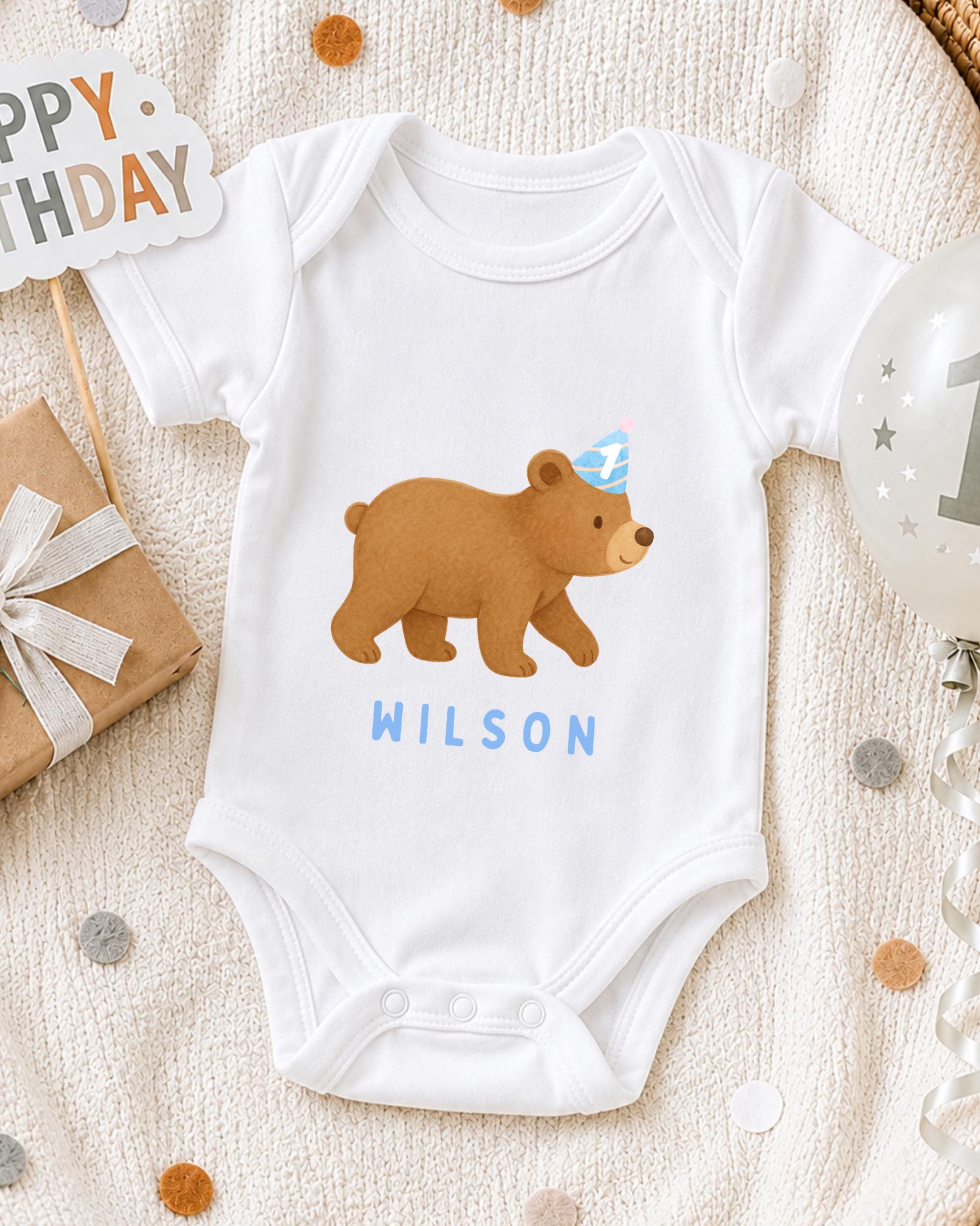 A white onesie with a bear design and the name "WILSON" printed on it, along with a birthday sign and a balloon, all arranged on a white blanket.