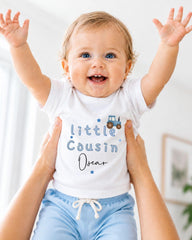 A smiling baby wearing a white shirt with the text "Little Cousin Oscar" is being held up by someone's arms.