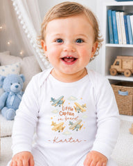 A smiling baby wearing a white onesie with a design of airplanes and the text "Little Captain on board" is sitting in a room with bookshelves and a teddy bear.