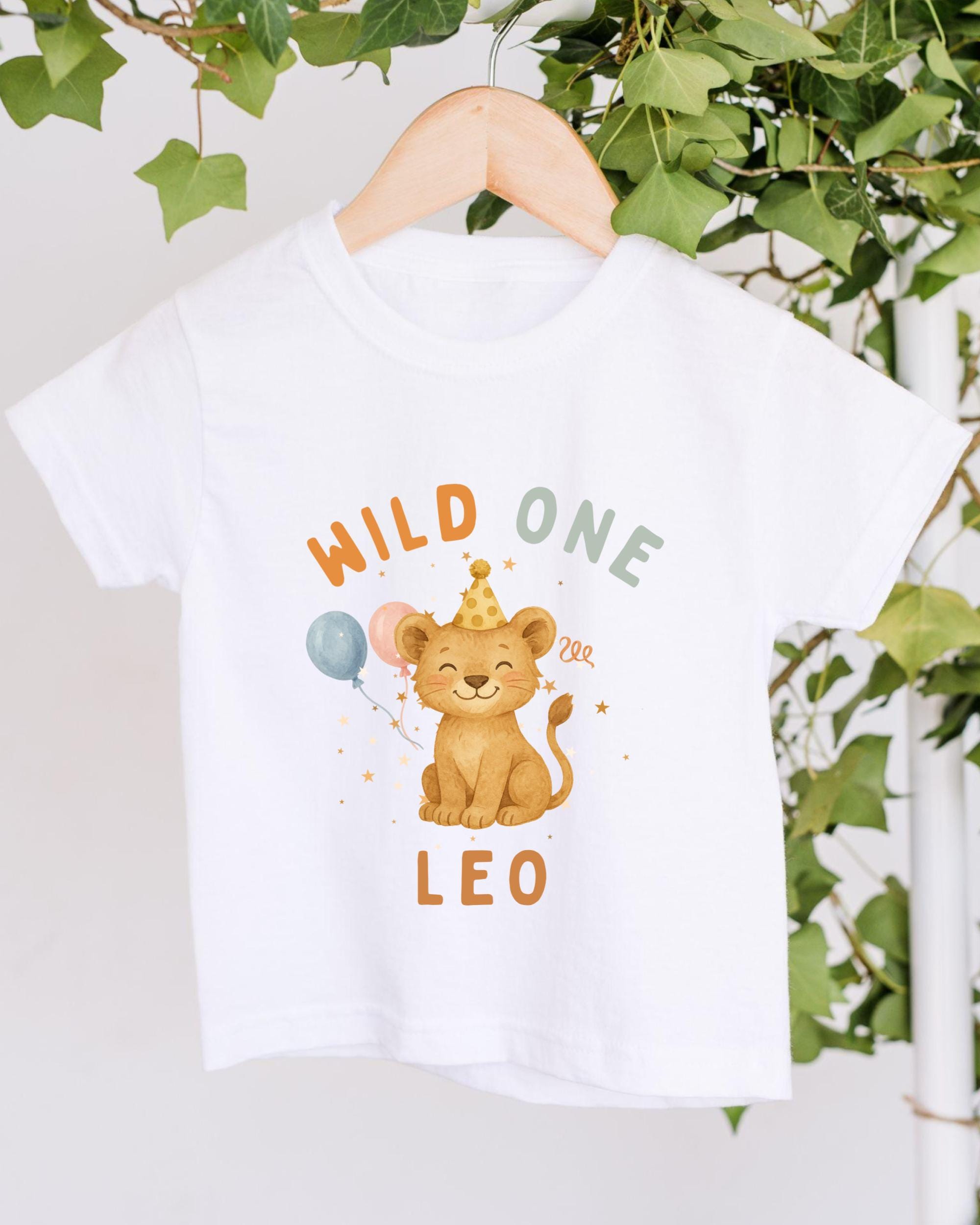 A white t-shirt with a cartoon image of a lion wearing a party hat and holding balloons, along with the text "Wild One Leo" printed on it.
