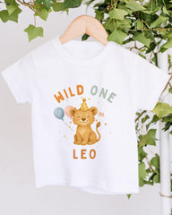 A white t-shirt with a cartoon image of a lion wearing a party hat and holding balloons, along with the text "Wild One Leo" printed on it.