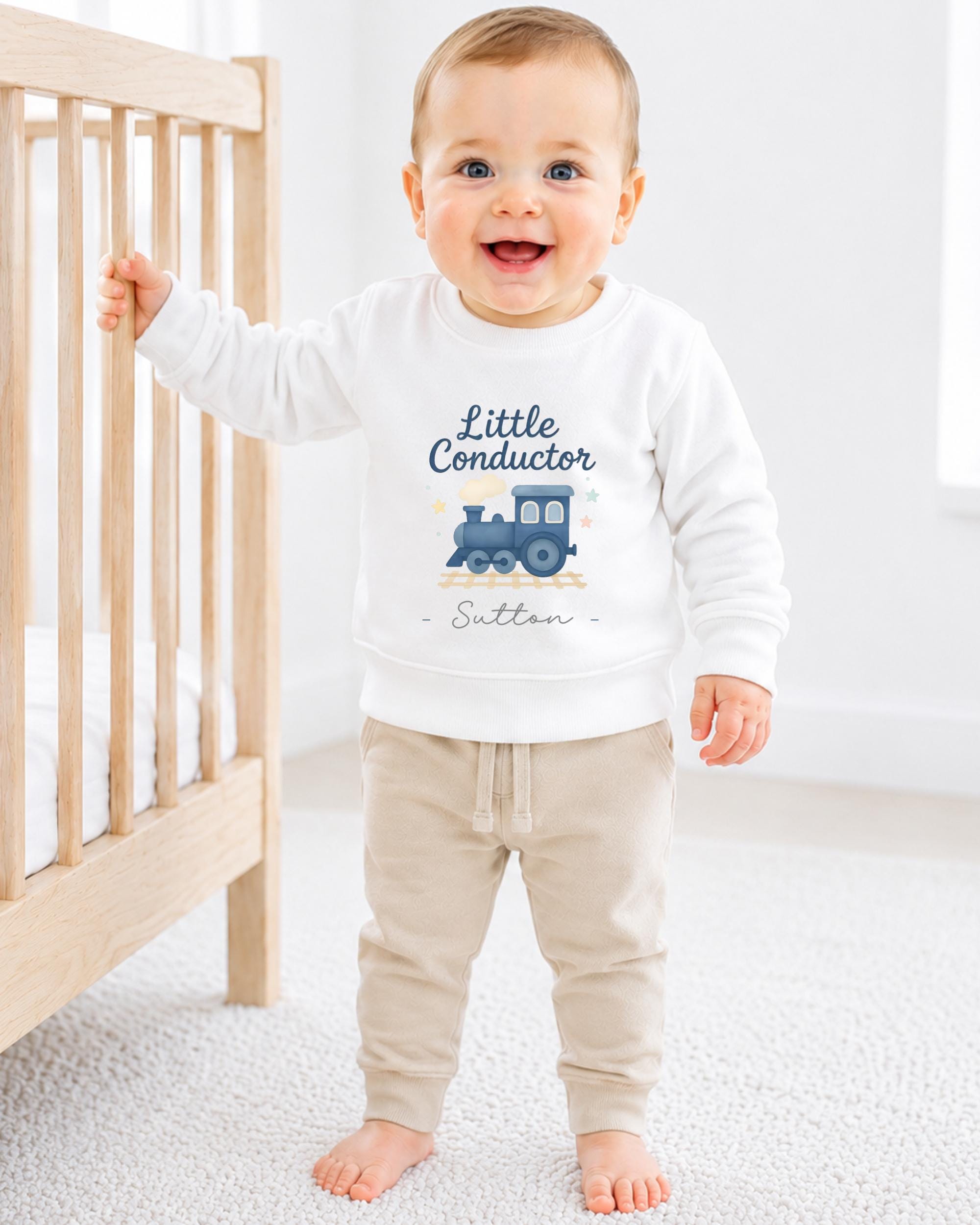 A smiling baby boy wearing a white sweatshirt with a train graphic and the text "Little Conductor" is standing next to a wooden crib.