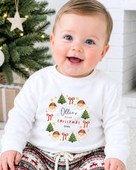 A smiling baby boy wearing a white sweatshirt with a Christmas wreath design and text on it, sitting in front of a Christmas tree.