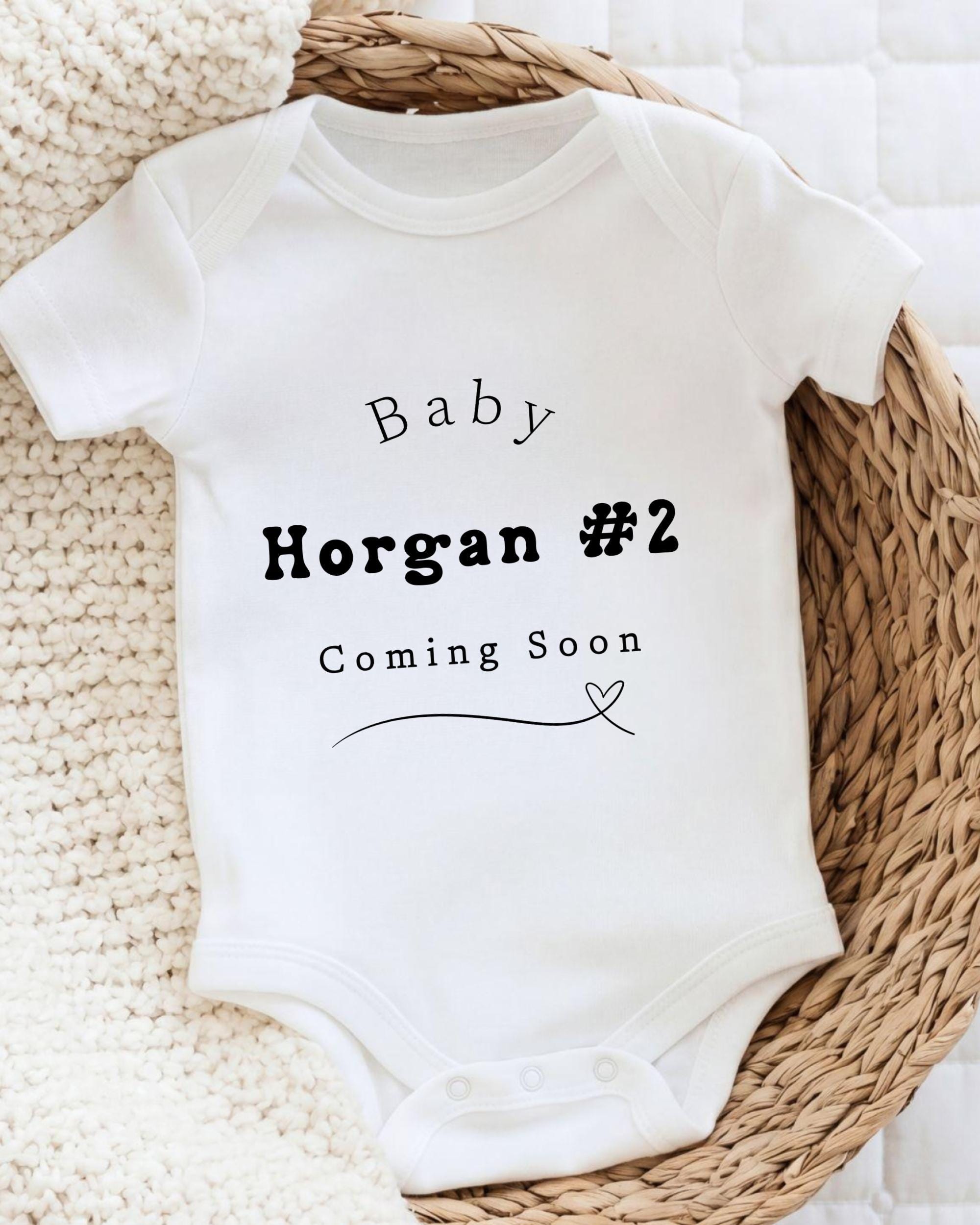 A white baby onesie with text that says "Baby Morgan #2 Coming Soon" printed on it.