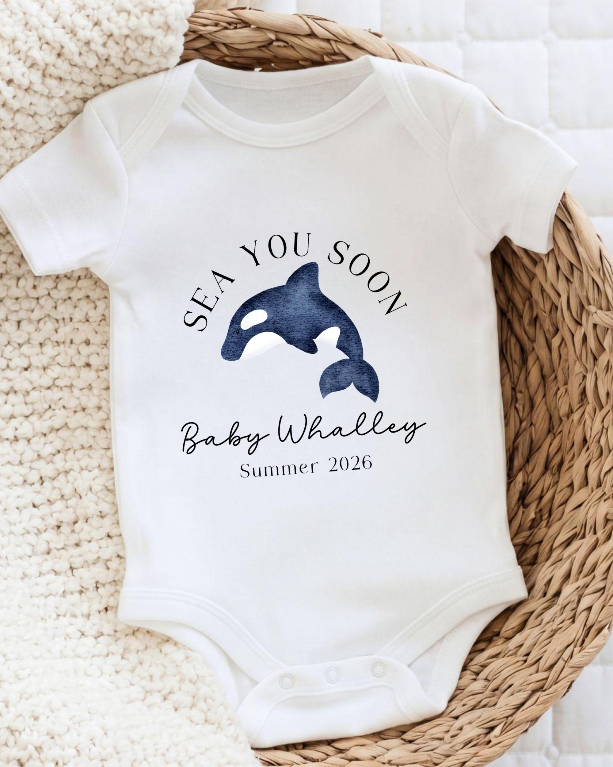 A white baby onesie with a graphic of a whale and the text "SEA YOU SOON Baby Whalley Summer 2026" printed on it.