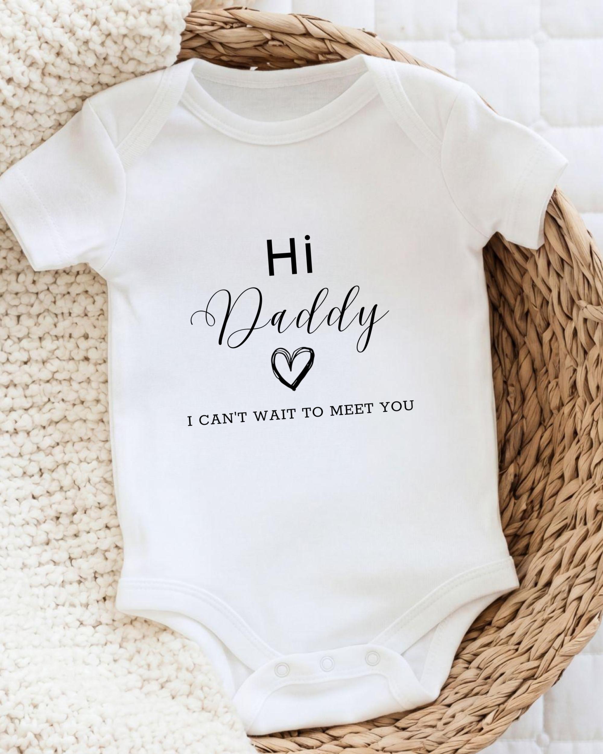 A white baby onesie with the text "Hi Daddy I can't wait to meet you" printed on it, placed on a woven basket.