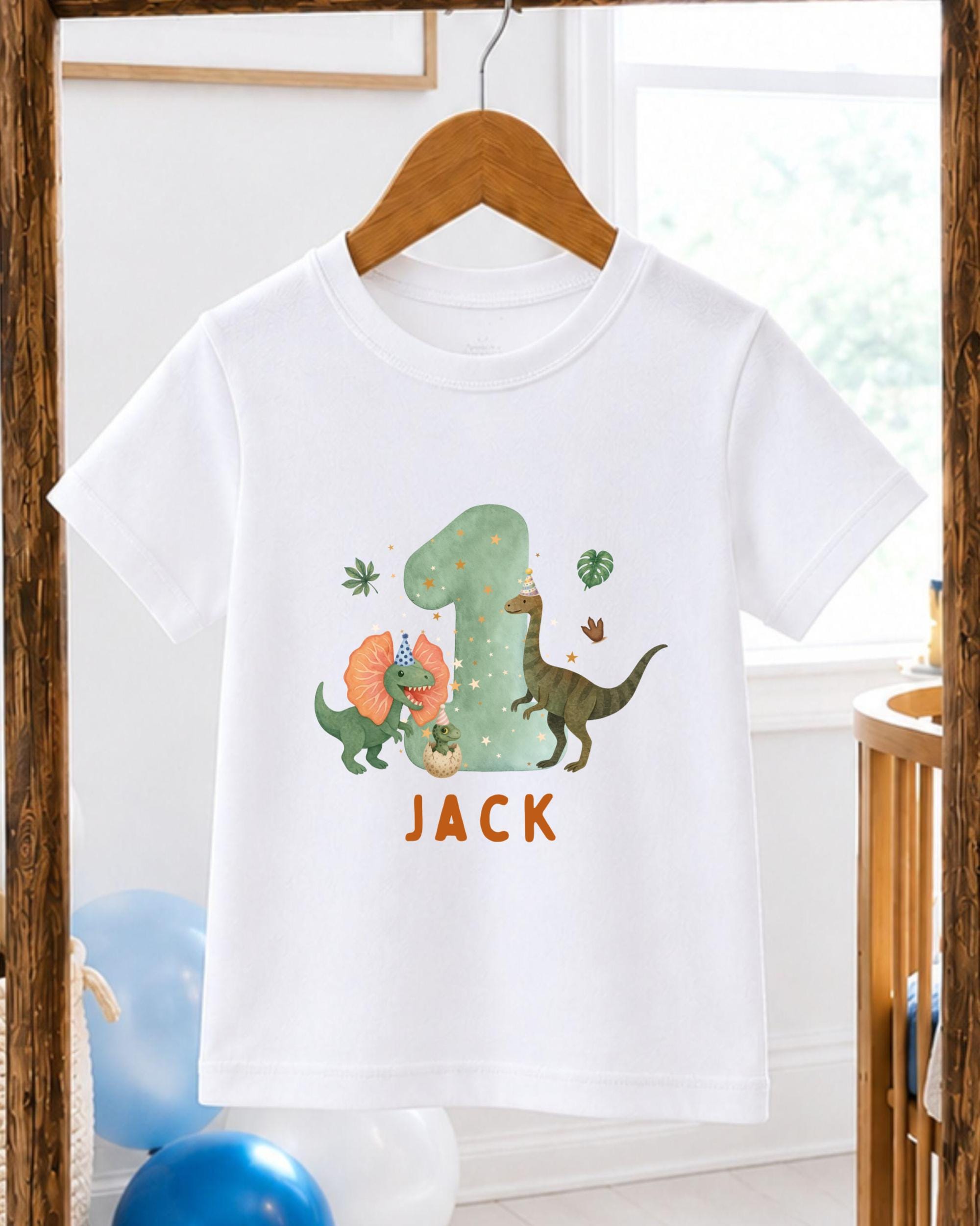 A white t-shirt with a dinosaur-themed design and the name "Jack" printed on it, hanging on a wooden hanger.
