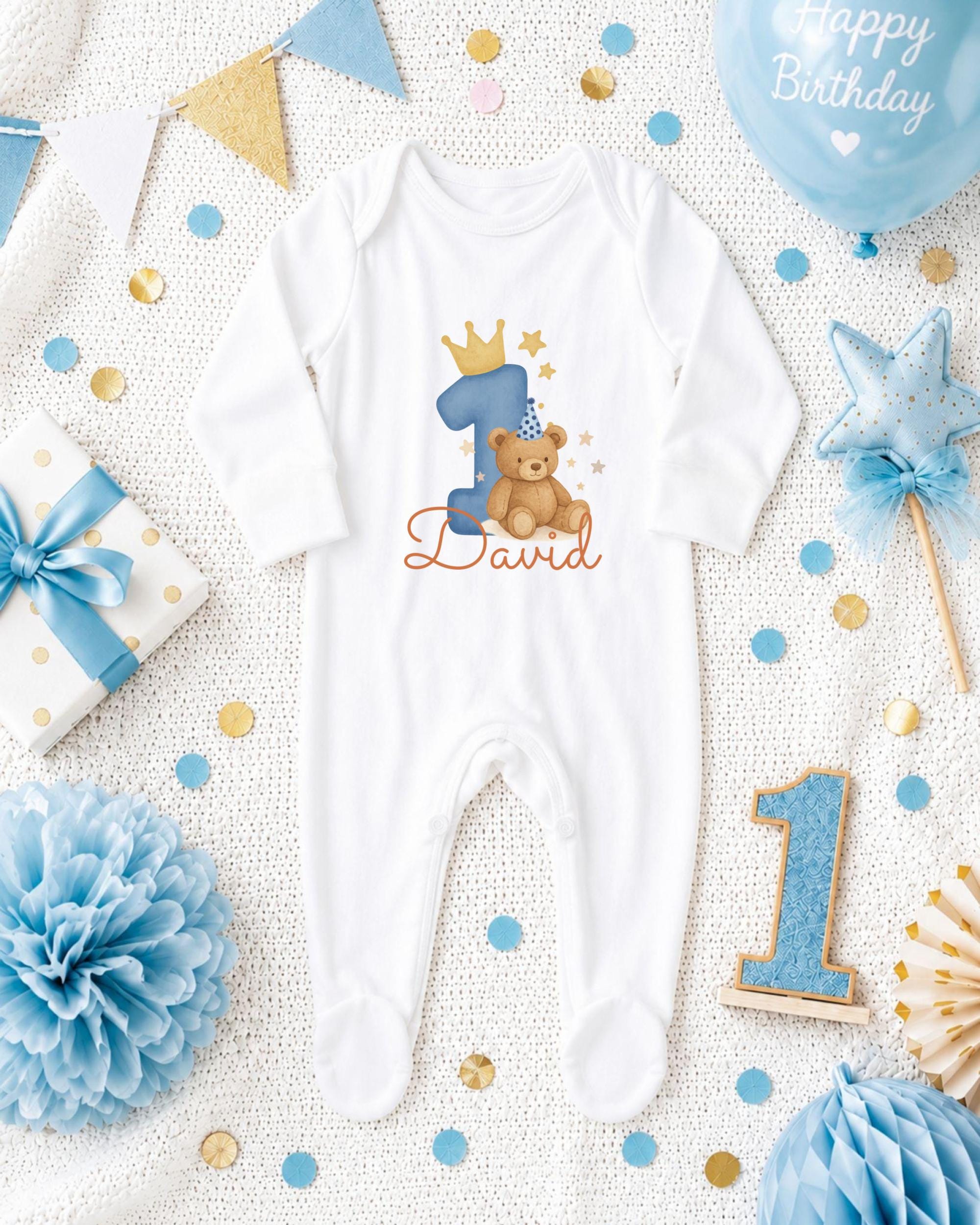 A white onesie with a teddy bear and the number 1, surrounded by birthday decorations such as a blue balloon, blue and gold confetti, and a blue and gold bow.