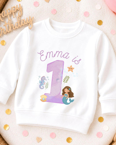 A white sweatshirt with a mermaid and a number 1 printed on it, surrounded by confetti.