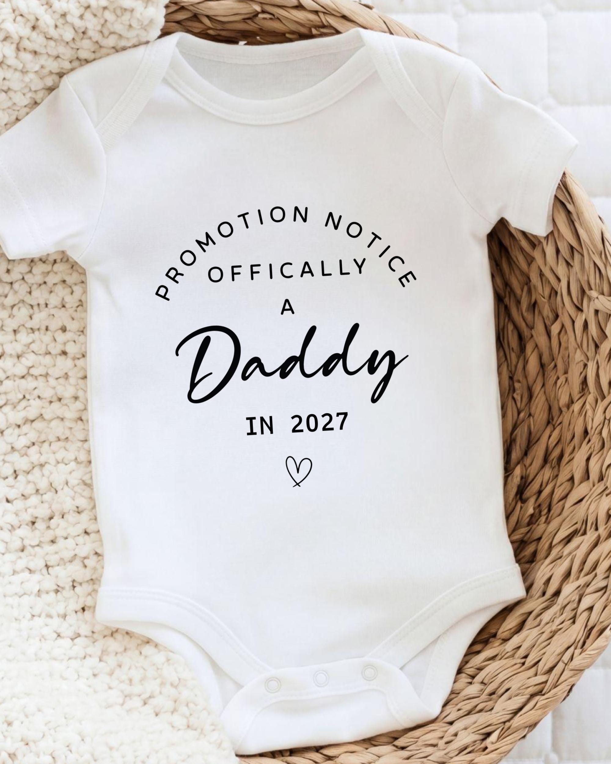 A white baby onesie with text printed on it, placed on a woven basket.