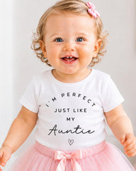 A smiling baby girl wearing a white t-shirt with text and a pink bow, standing in front of a white background.