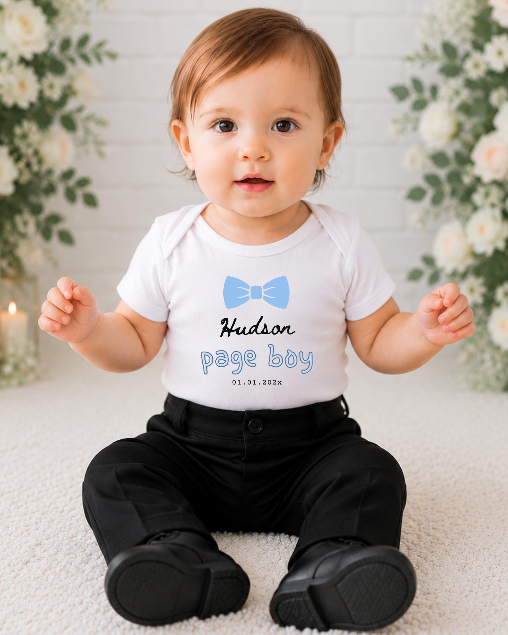 A smiling baby boy wearing a white shirt with a blue bow tie and black pants sits on a white carpet, with a floral background behind him.