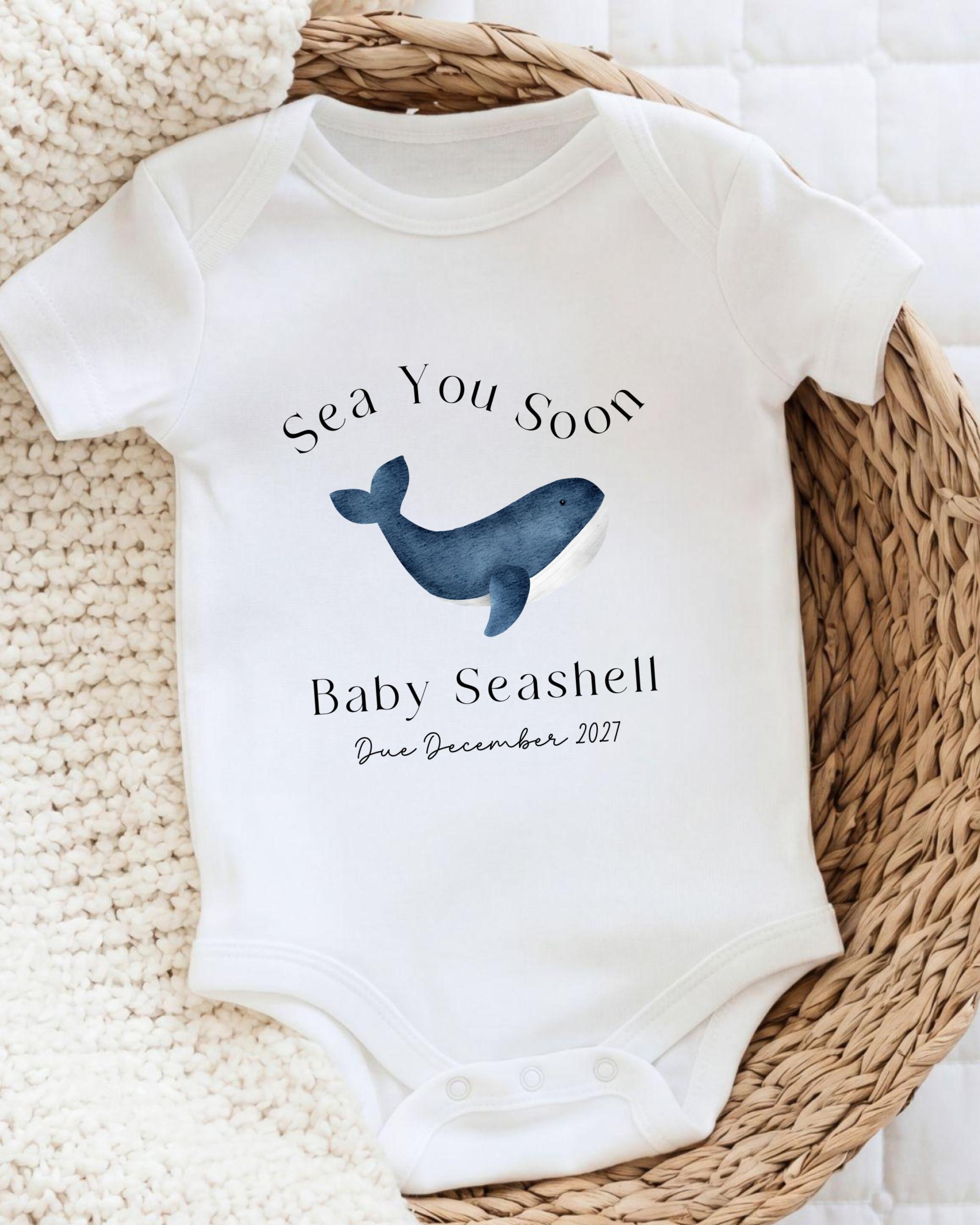 A white baby onesie with a blue whale graphic and text that reads "Sea You Soon Baby Seashell Due December 2021".