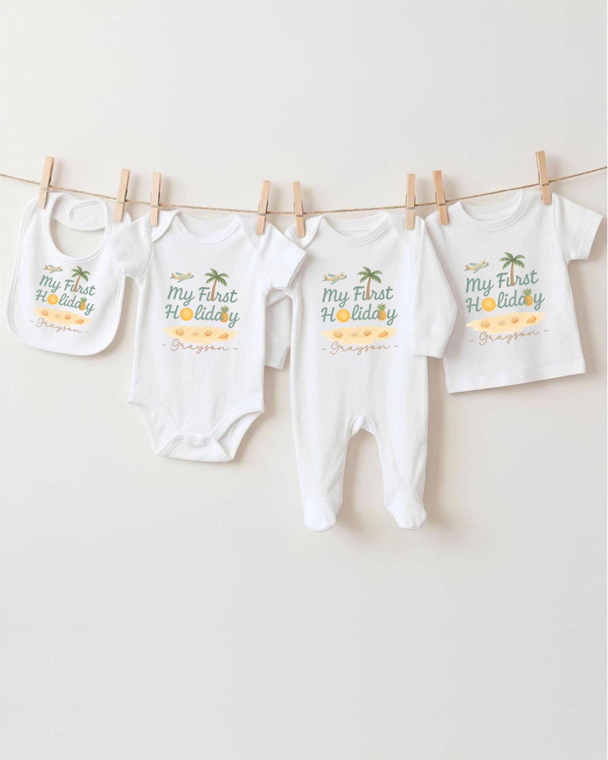 A row of white baby clothes with the text "My first holiday" printed on them, hanging on a clothesline.