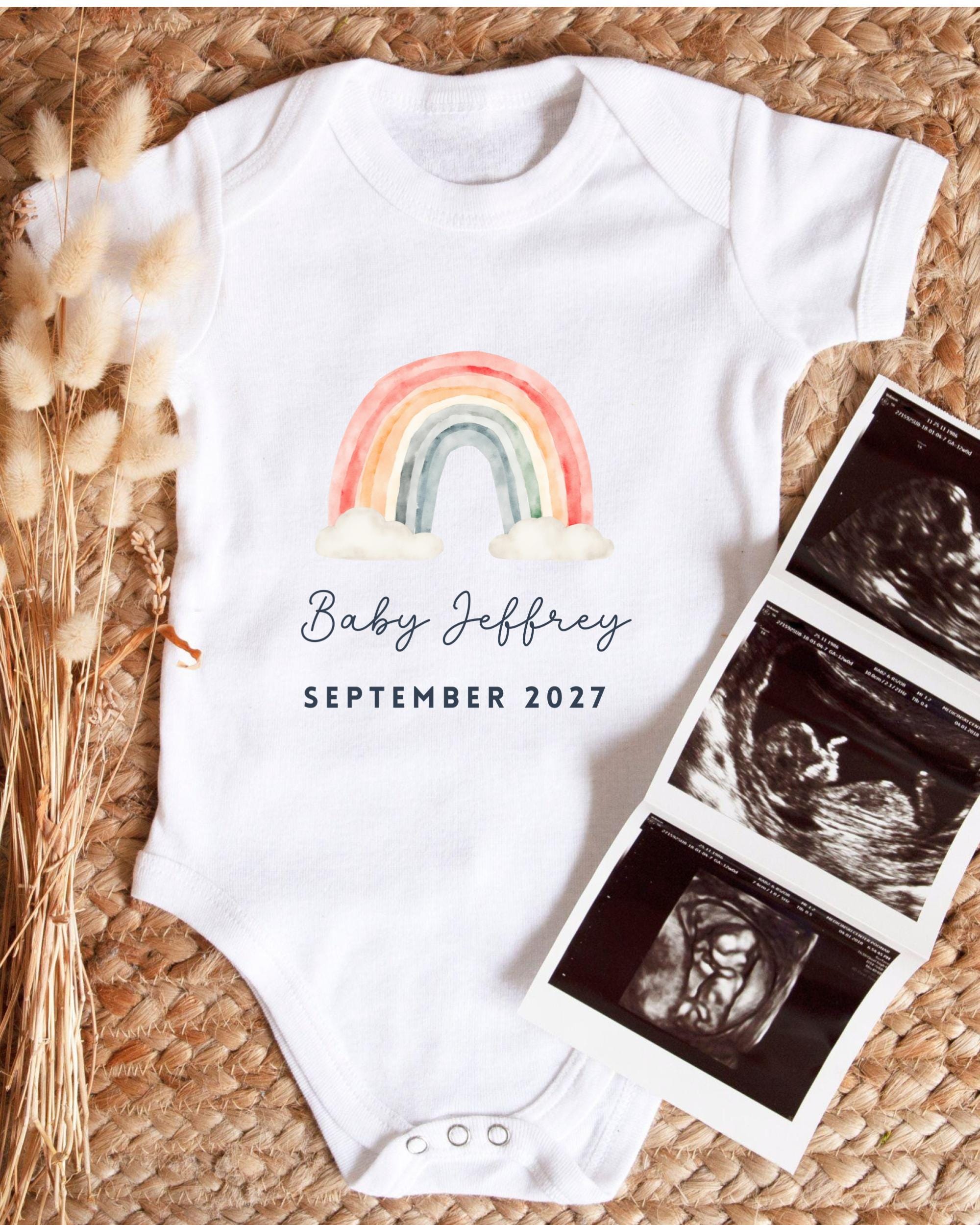 A white baby onesie with a rainbow graphic and the text "Baby Jeffrey September 2027" printed on it, along with a photograph of an ultrasound image.
