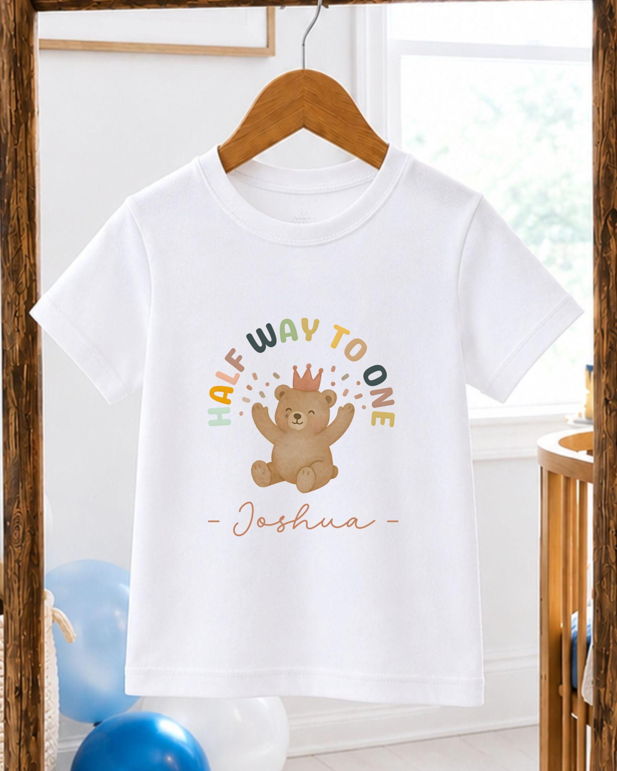 A white t-shirt with a colorful graphic of a teddy bear wearing a crown, along with the text "Half Way to One - Joshua" printed on it.