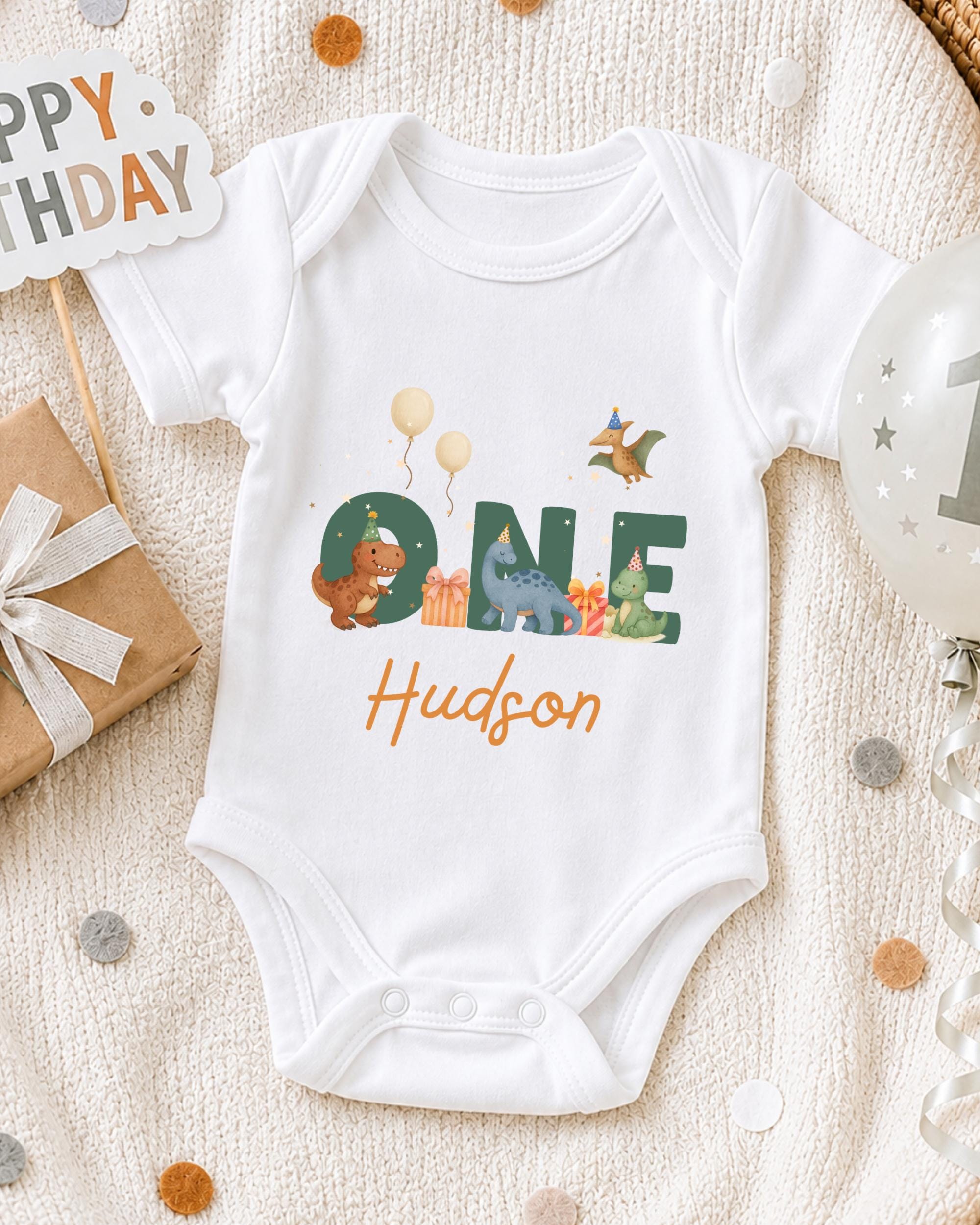 A white onesie with the name "Hudson" printed on it, surrounded by various birthday decorations such as balloons, a gift box, and confetti.