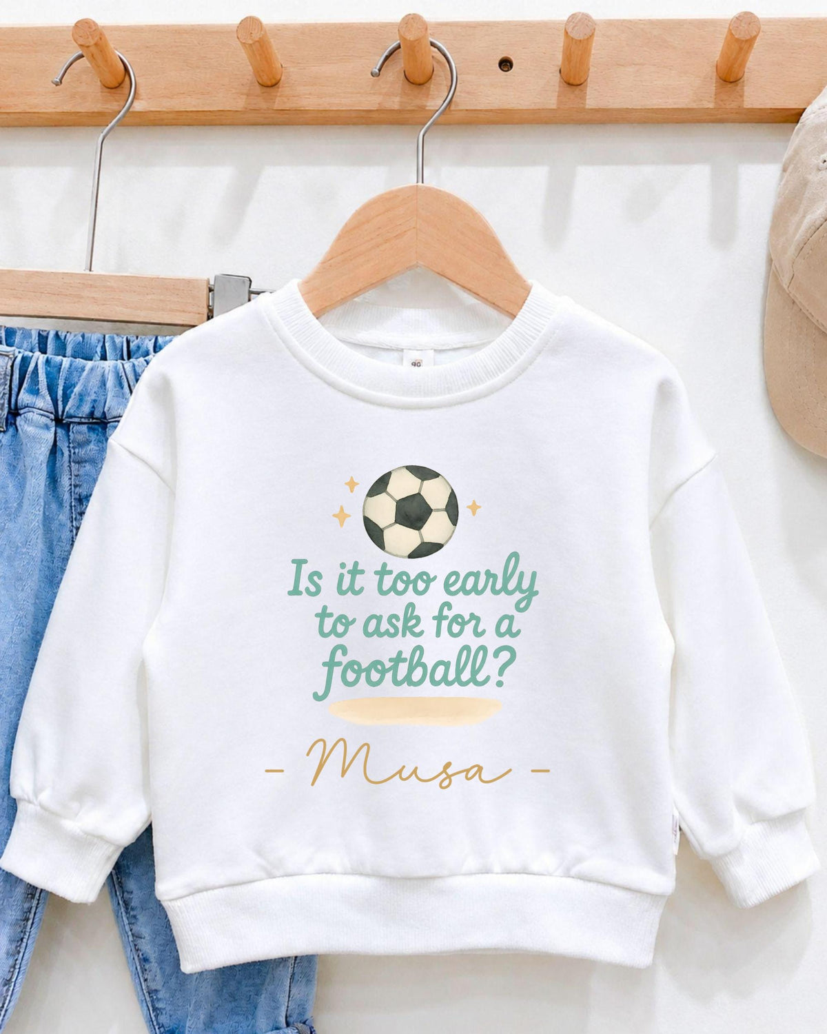 A white sweatshirt with a soccer ball design and text printed on it, hanging on a wooden hanger.