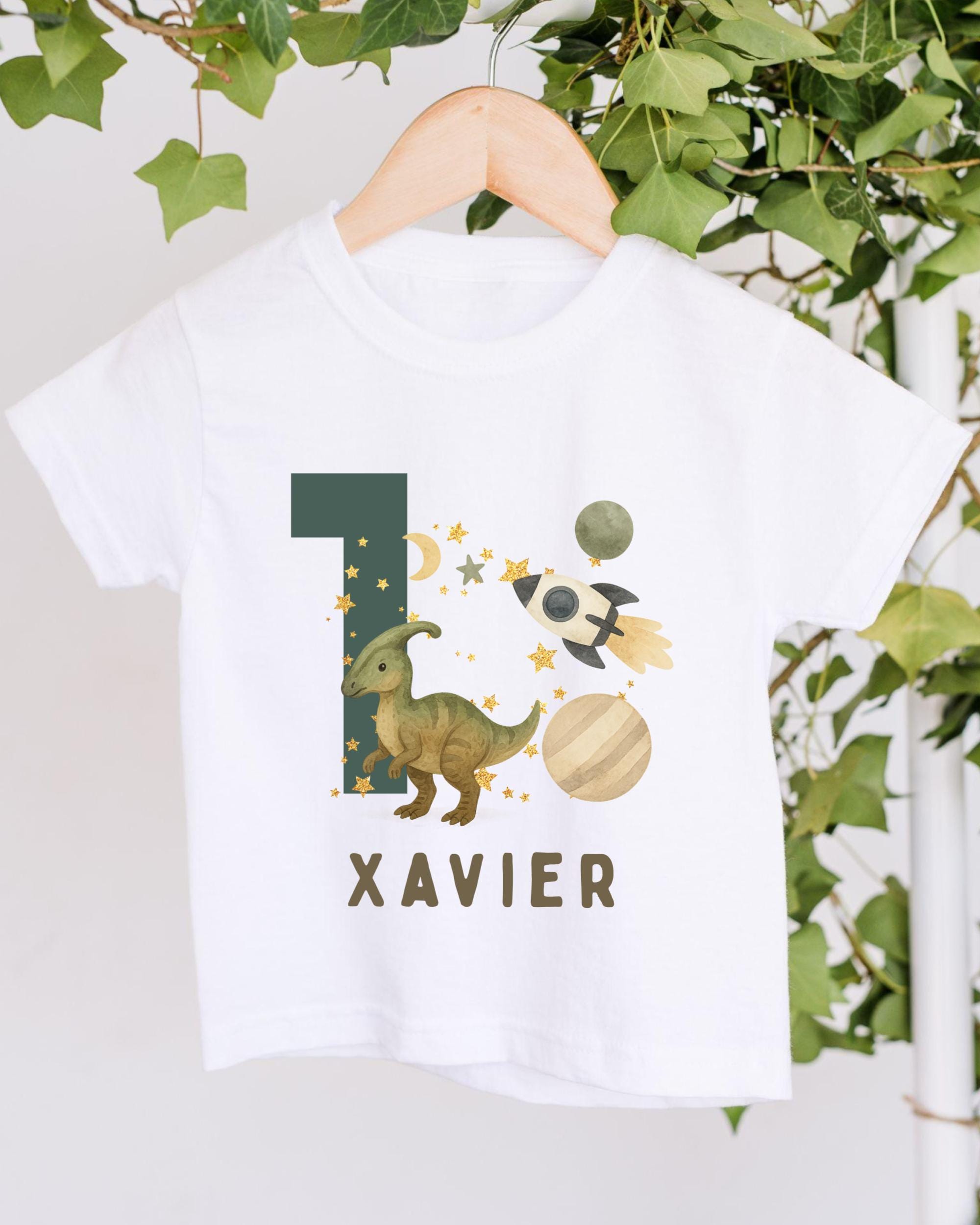 A white t-shirt with a dinosaur graphic and the number 1, along with a rocket ship and stars, hanging on a wooden hanger.
