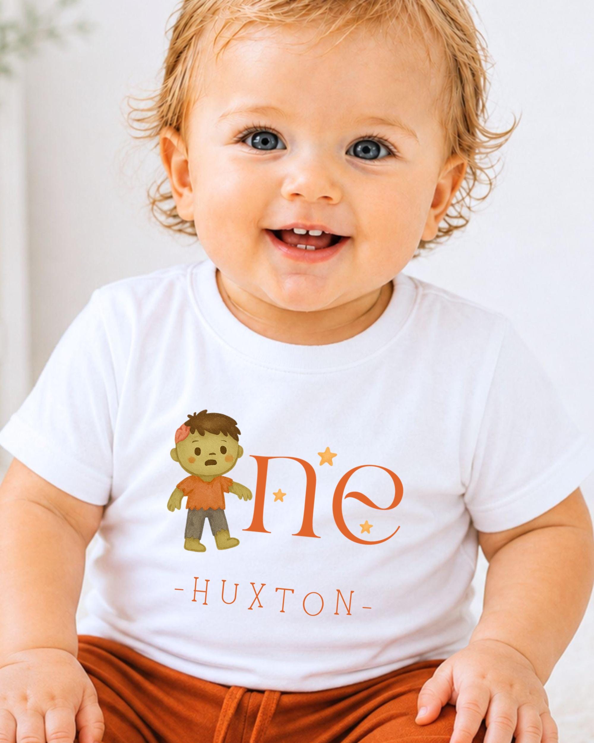 A smiling baby wearing a white t-shirt with a cartoon character and the text "One - Huxton" printed on it.