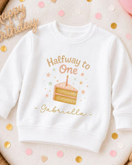 A white sweatshirt with a birthday cake design and the text "Halfway to One Gabriella" printed on it, surrounded by confetti.