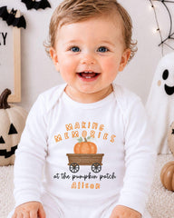 A smiling baby wearing a white onesie with a pumpkin and carriage graphic, sitting in front of Halloween decorations.