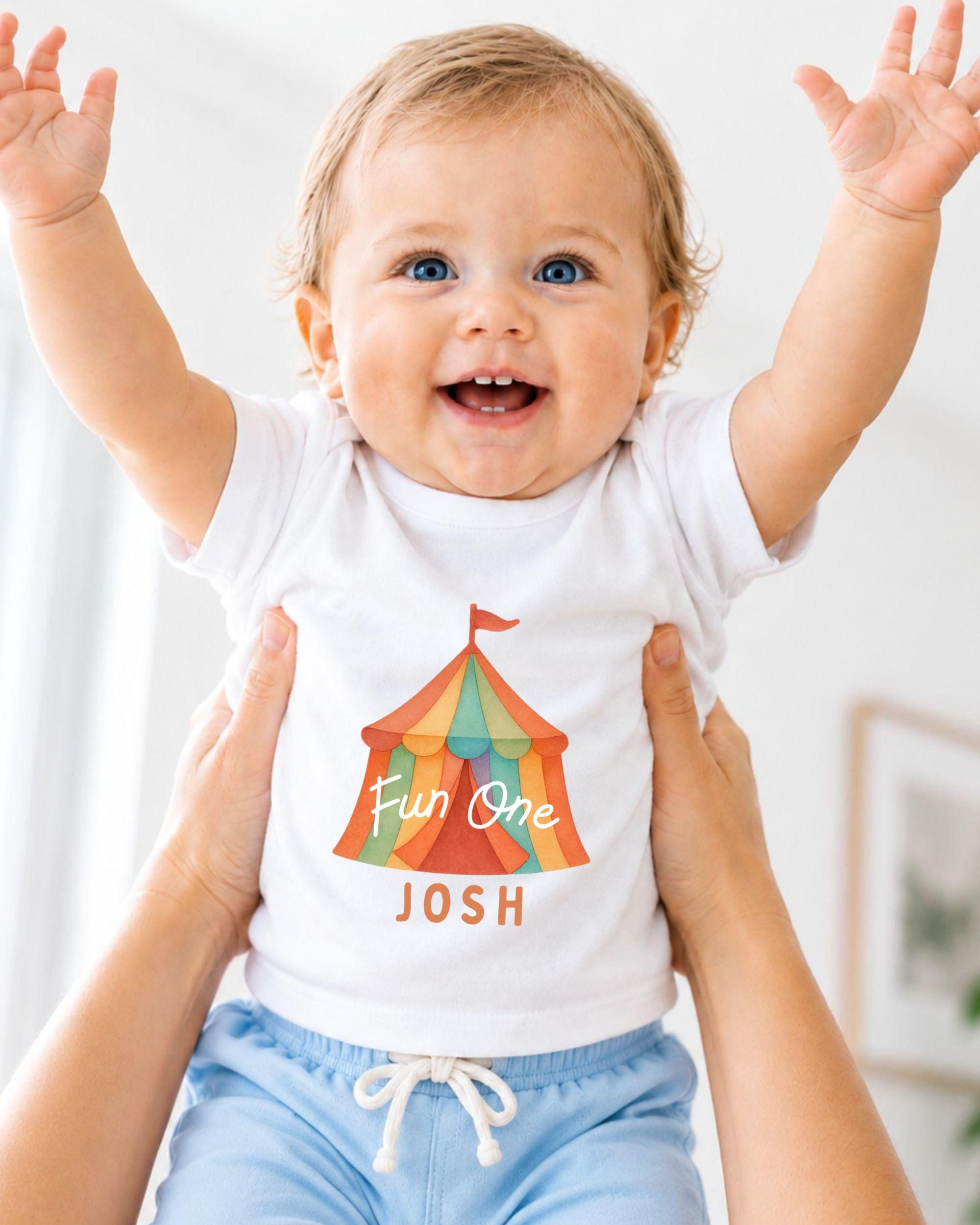 A smiling baby wearing a white t-shirt with a colorful circus tent graphic and the name " Josh" printed on it, being held up by someone's arms.